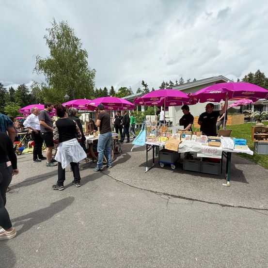 Ein Outdoor-Markt mit mehreren Ständen unter lila Schirmen. Mehrere Leute stehen herum und unterhalten sich. Ein Mann ordnet Gegenstände auf einem Tisch an.