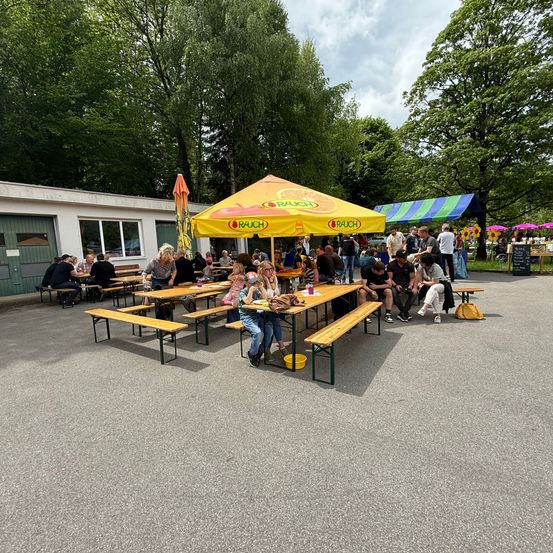 Im Freien essen mit einer großen Gruppe von Menschen, die unter einem Schirm sitzen, umgeben von Bäumen und Bänken.