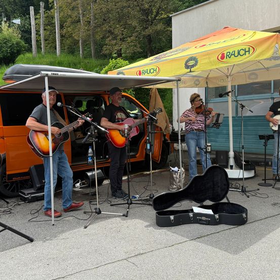 Eine Band spielt Musik im Freien mit einem orangefarbenen Van im Hintergrund. Zwei Männer spielen Gitarre, eine Frau spielt Geige und ein Etui liegt auf dem Boden.