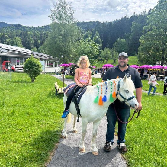 Ein Mann steht neben einem kleinen Kind auf einem weißen Pferd in einem Park mit grünem Gras und Bäumen, mit einem Gebäude und einem roten Auto im Hintergrund.