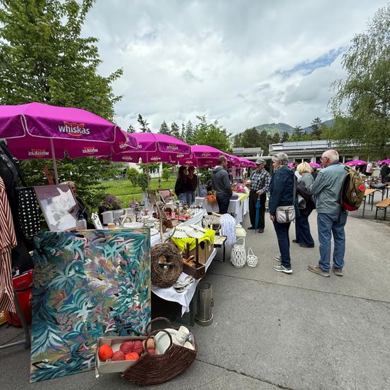 Outdoor-Markt mit lila Regenschirmen. Gegenstände sind auf Tischen ausgestellt. Leute stöbern durch die Artikel. Ein großes Gemälde liegt auf dem Boden.