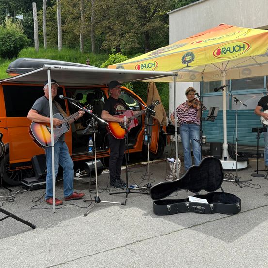 Eine Gruppe von Musikern spielt Gitarren und eine Geige unter einem großen Regenschirm vor einem Gebäude und einem orangefarbenen Van.