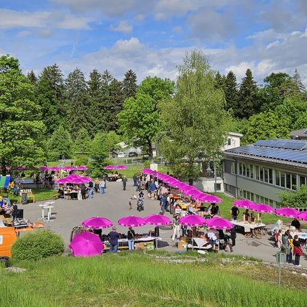 Ein Freiluftmarkt mit vielen lilafarbenen Schirmen, einkaufende Menschen und ein Gebäude mit Solarmodulen im Hintergrund.