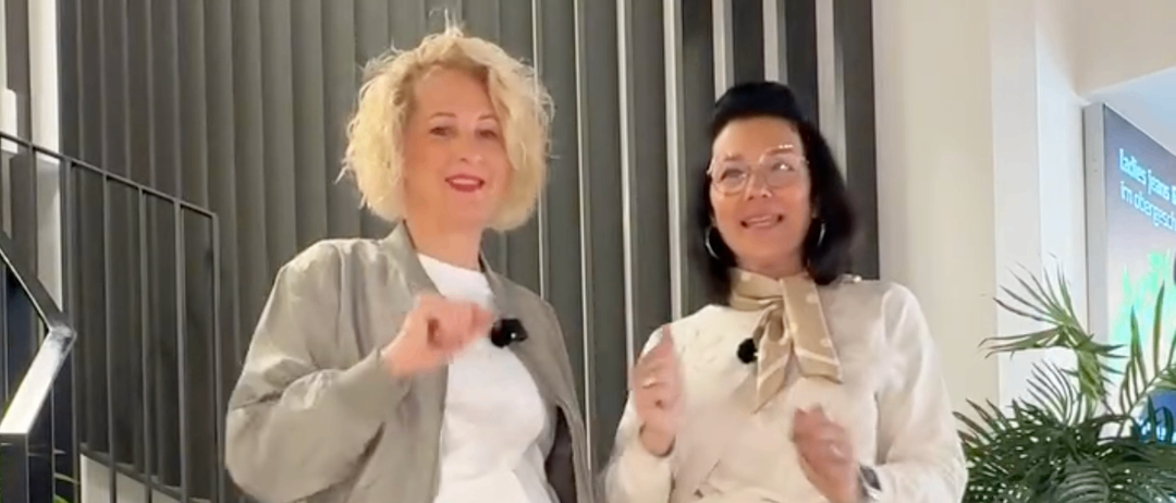 Zwei Frauen stehen auf einer Treppe, eine trägt eine beige Jacke und Hose, die andere ein weißes Oberteil und eine beige Rock. Beide lächeln und gestikulieren. Hinter ihnen steht eine Topfpflanze und ein Schild mit fremdsprachigem Text.