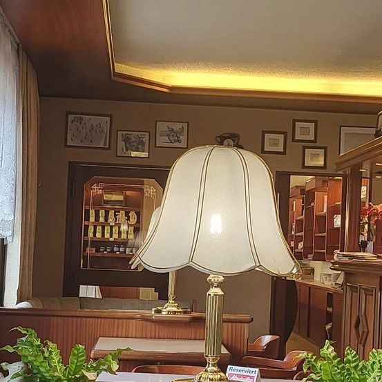 Ein Raum mit einem weißen Lampenschirm auf einem Tisch, umgeben von Holzmöbeln. Bilderrahmen schmücken die Wand, und ein Schrank ist im Hintergrund zu sehen.