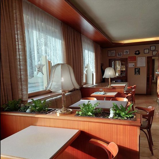 Ein Restaurantinnenraum mit Holztischen und Stühlen, Lampen und Pflanzen. Der Raum hat Vorhänge, eine weiße Decke und Bilderrahmen an der Wand.