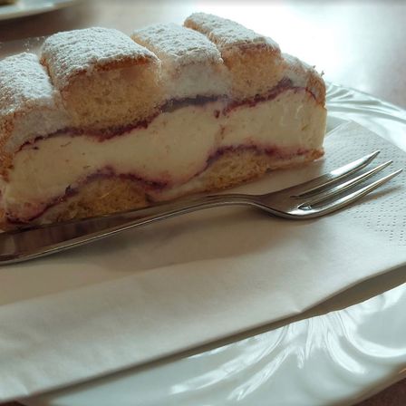 Ein Stück Kuchen mit einer cremigen Füllung und einem Hauch roter Früchte auf einem weißen Teller, begleitet von einer silbernen Gabel.