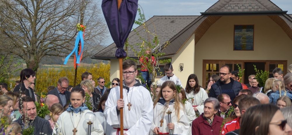 Eine Gruppe von Menschen in weißen Roben steht vor einem Gebäude und hält Kerzen. Der Mann in der Mitte trägt eine Brille und einen violetten Umhang. Sie sind von anderen Menschen umgeben, von denen einige ebenfalls eine Brille tragen.