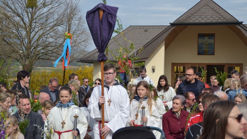 Eine Gruppe von Menschen in weißen Roben steht vor einem Gebäude und hält Kerzen. Der Mann in der Mitte trägt eine Brille und einen violetten Umhang. Sie sind von anderen Menschen umgeben, von denen einige ebenfalls eine Brille tragen.