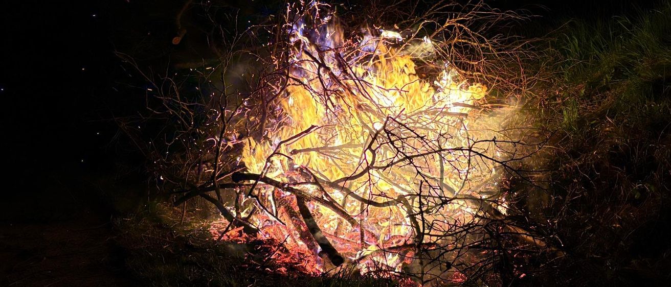 Ein großes Feuer brennt in der Dunkelheit, umgeben von Ästen und Zweigen, wobei Funken in die Luft fliegen.