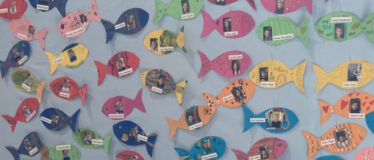 Ein blauer Hintergrund mit vielen Fischbildern, jeweils mit einem Namen beschriftet, ist ausgestellt. Darunter steht ein Tisch mit vielen kleinen Flaschen in Keramikschuhen, einige mit einem Kreuzsymbol.