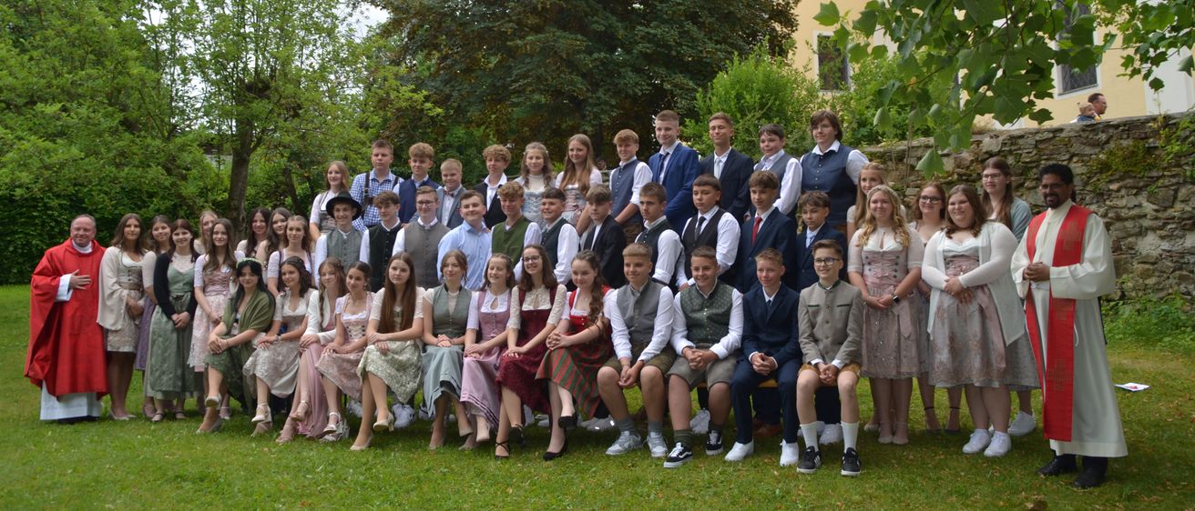 Eine Gruppe von Schülern posiert für ein Foto im Freien. Sie sind in traditioneller Kleidung gekleidet, einige in weißen Hemden und andere in grünen und blauen Westen. Der Hintergrund besteht aus einer Mischung aus grünen Bäumen und einer Steinmauer.