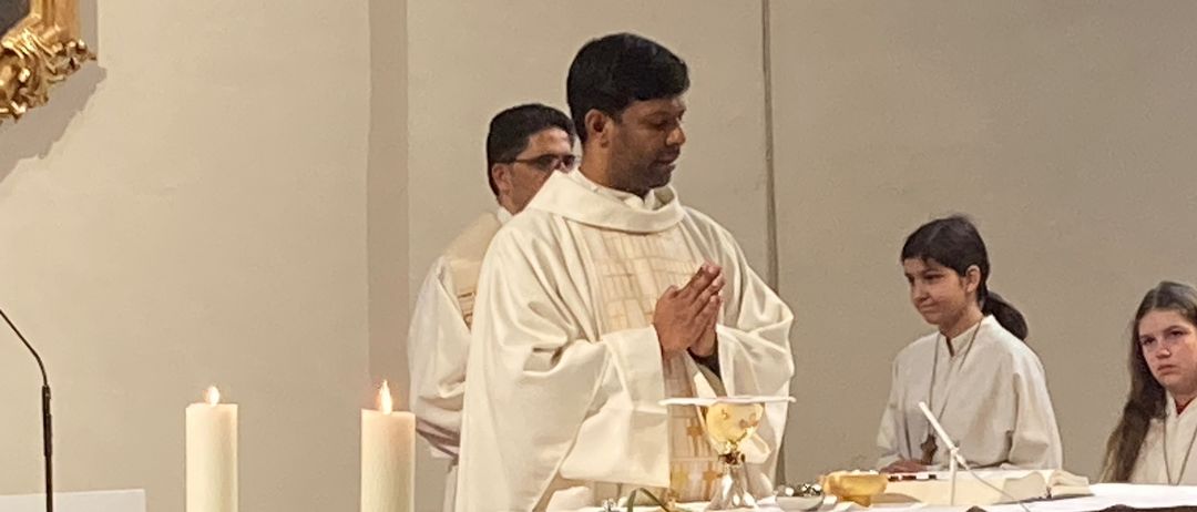 Ein Priester betet vor dem Altar mit zwei brennenden Kerzen, während eine Frau einen Kelch hält. Ein anderer Priester steht hinter ihm.