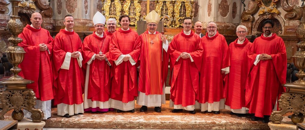 Eine Gruppe von Priestern in roten Roben steht vor einem Altar mit goldenen Kerzenhaltern.