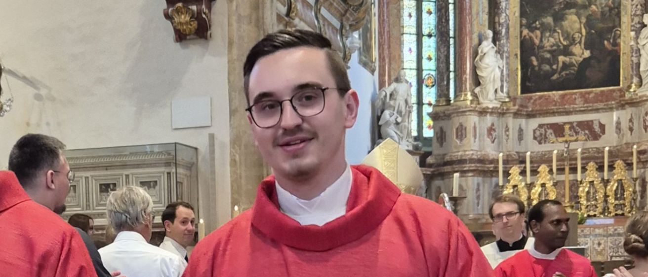 Zwei Männer in roten Gewändern, wahrscheinlich Priester, stehen Seite an Seite in einer Kirche mit gefalteten Händen vor sich. Hinter ihnen stehen andere Menschen und beobachten.