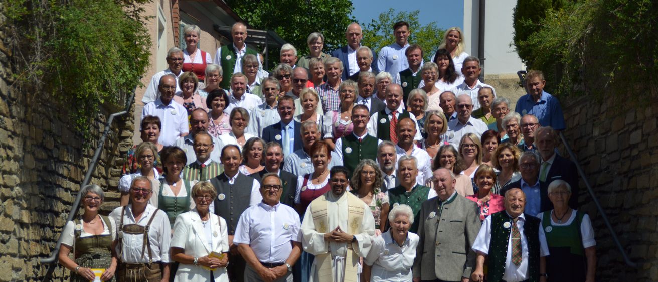 Eine große Gruppe älterer Menschen, einige in traditioneller Kleidung, versammelt sich vor einem Gebäude, mit einem Priester in ihrer Mitte.