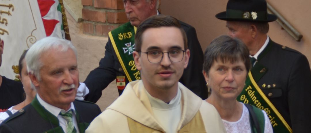 Eine Gruppe von Menschen steht in einer Reihe draußen. Ein junger Mann in einem goldenen Gewand steht in der Mitte, während mehrere Menschen in traditioneller deutscher Kleidung hinter ihm stehen. Ein Fahnenmast und eine Treppe sind im Hintergrund zu sehen.