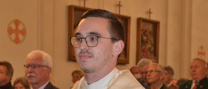 Ein Priester steht in einer Kirche und trägt eine goldene Stola. Hinter ihm beobachten Männer in Anzügen und Krawatten, darunter einer mit roter Krawatte.