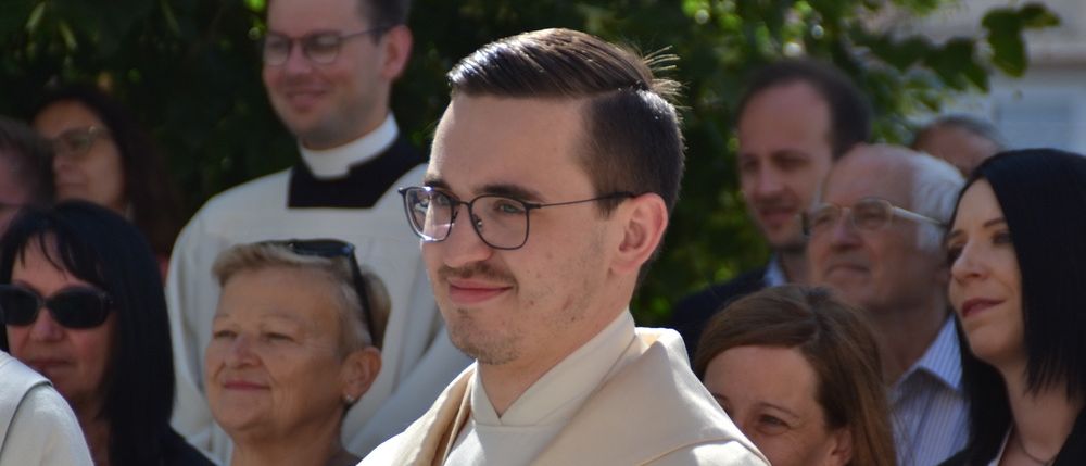 Ein Priester in einer weißen und goldenen Robe steht vor einer Gruppe von Menschen, von denen einige eine Brille tragen.