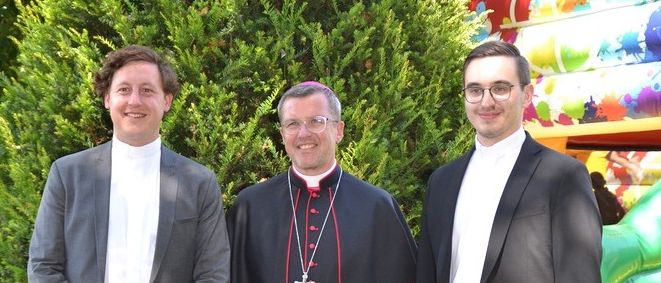 Drei Männer stehen vor einer Hecke mit einem aufblasbaren Sprungschloss dahinter. Zwei tragen Anzüge und einer ist als Bischof gekleidet.