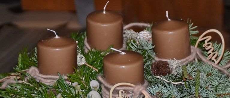 Ein Kranz mit vier braunen Kerzen und Tannennadeln, verziert mit zwei Holzschildern, die 'Fried' und 'Liebe' sagen. Er steht auf einem Holztisch.