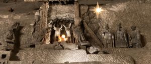 Eine Krippe aus Stein ist in einer Höhle ausgestellt, mit Figuren von Maria, Josef und Jesus sowie Tieren. Ein heller Stern hängt darüber und die Szene wird von einem kleinen Feuer beleuchtet.