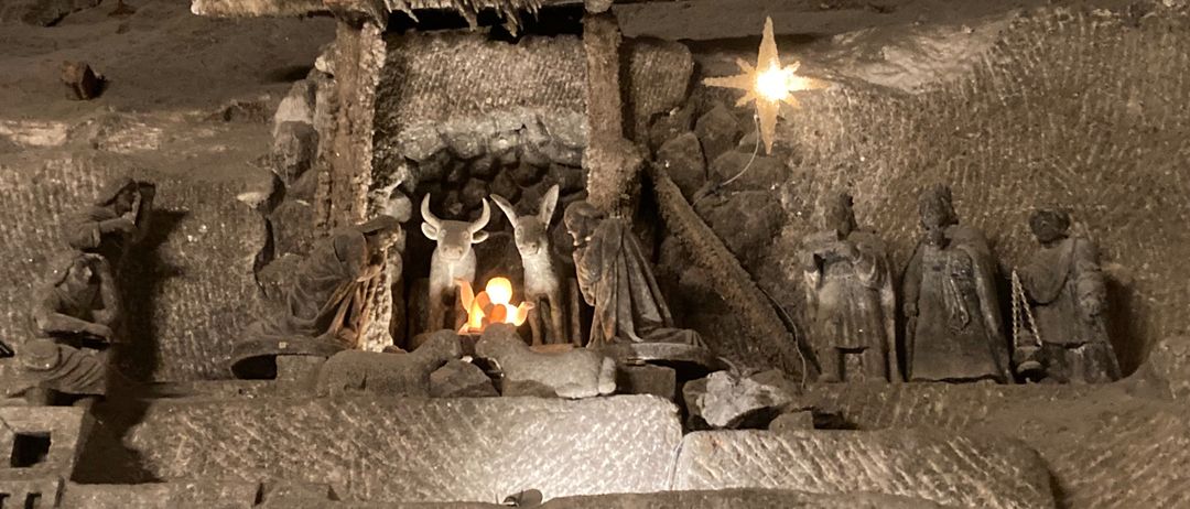 Eine Krippe aus Stein ist in einer Höhle ausgestellt, mit Figuren von Maria, Josef und Jesus sowie Tieren. Ein heller Stern hängt darüber und die Szene wird von einem kleinen Feuer beleuchtet.