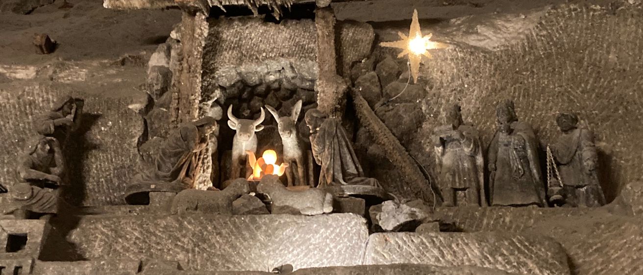 Eine Krippe aus Stein ist in einer Höhle ausgestellt, mit Figuren von Maria, Josef und Jesus sowie Tieren. Ein heller Stern hängt darüber und die Szene wird von einem kleinen Feuer beleuchtet.