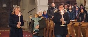 Eine Gruppe von Menschen, darunter zwei Frauen mit Kerzen, geht in einer Kirche. Ein Mädchen trägt eine Puppe. Der Raum hat gewölbte Decken, Holzbänke und eine Kristallleuchter.