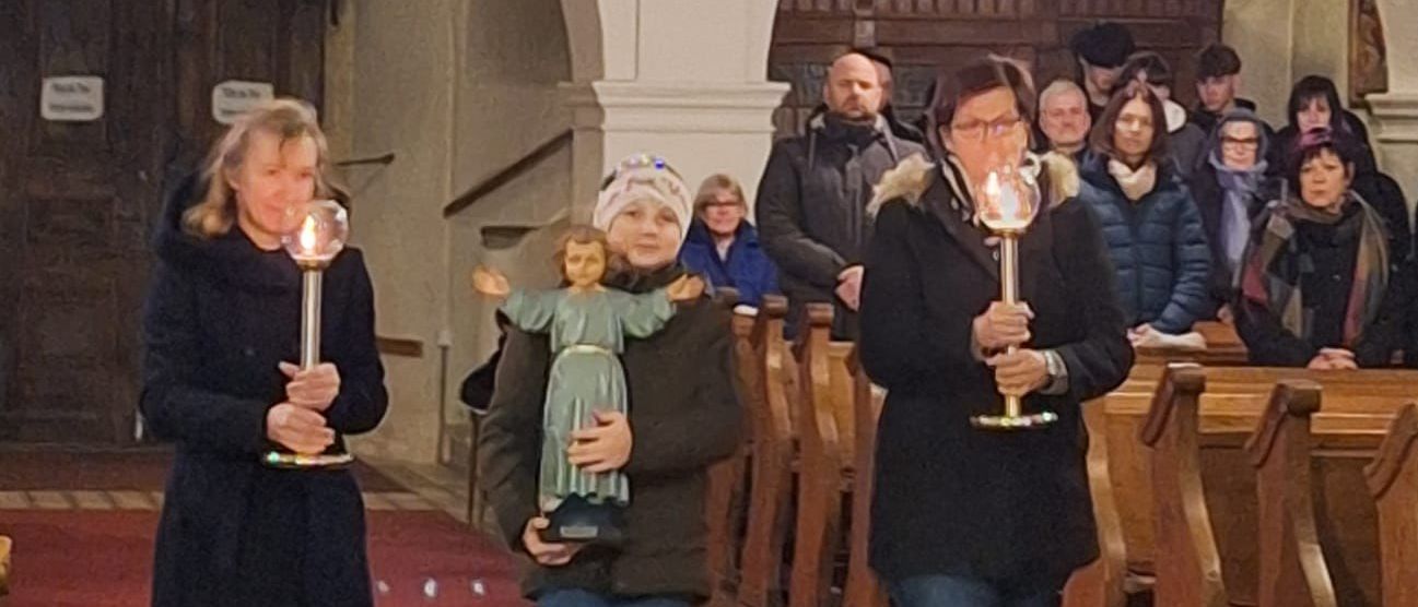 Eine Gruppe von Menschen, darunter zwei Frauen mit Kerzen, geht in einer Kirche. Ein Mädchen trägt eine Puppe. Der Raum hat gewölbte Decken, Holzbänke und eine Kristallleuchter.