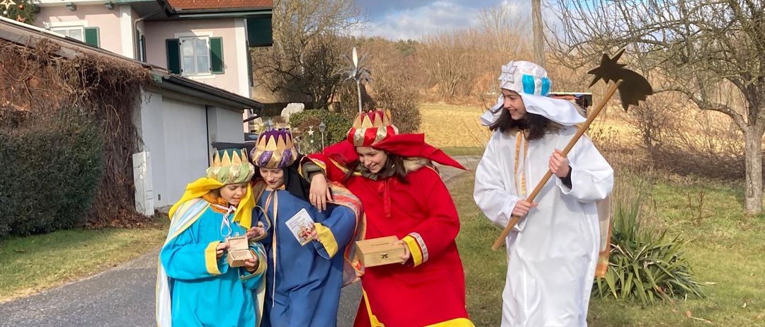 Fünf Personen in Kostümen stehen vor einem Haus. Zwei sind in Blau, einer in Rot und zwei in Weiß. Sie halten Requisiten und scheinen aufzutreten.