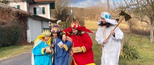 Fünf Personen in Kostümen stehen vor einem Haus. Zwei sind in Blau, einer in Rot und zwei in Weiß. Sie halten Requisiten und scheinen aufzutreten.