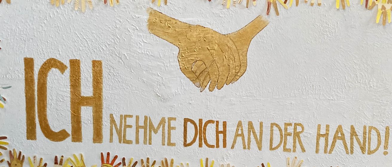 Ein großes Banner zeigt eine goldene Hand, die sich ausstreckt, mit den Worten 'Ich nehme dich an der Hand!' darüber, umgeben von vielen bunten Händen.