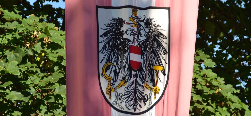 Eine Fahne mit einem roten und weißen Emblem, das einen Doppeladler, einen Hammer und ein Zepter zeigt, vor einem rosa und weißen Hintergrund mit grünen Blättern.