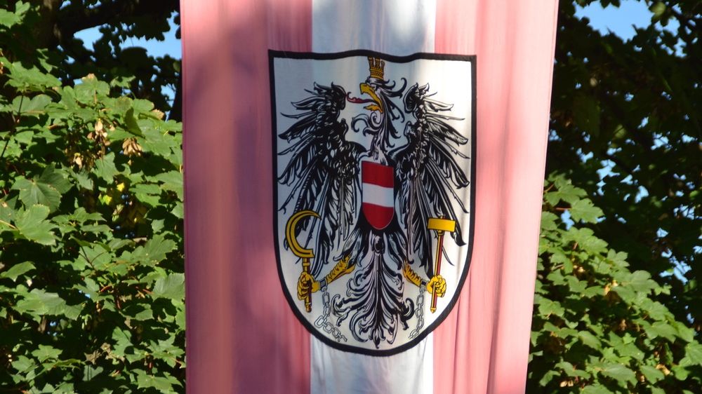 Eine Fahne mit einem roten und weißen Emblem, das einen Doppeladler, einen Hammer und ein Zepter zeigt, vor einem rosa und weißen Hintergrund mit grünen Blättern.