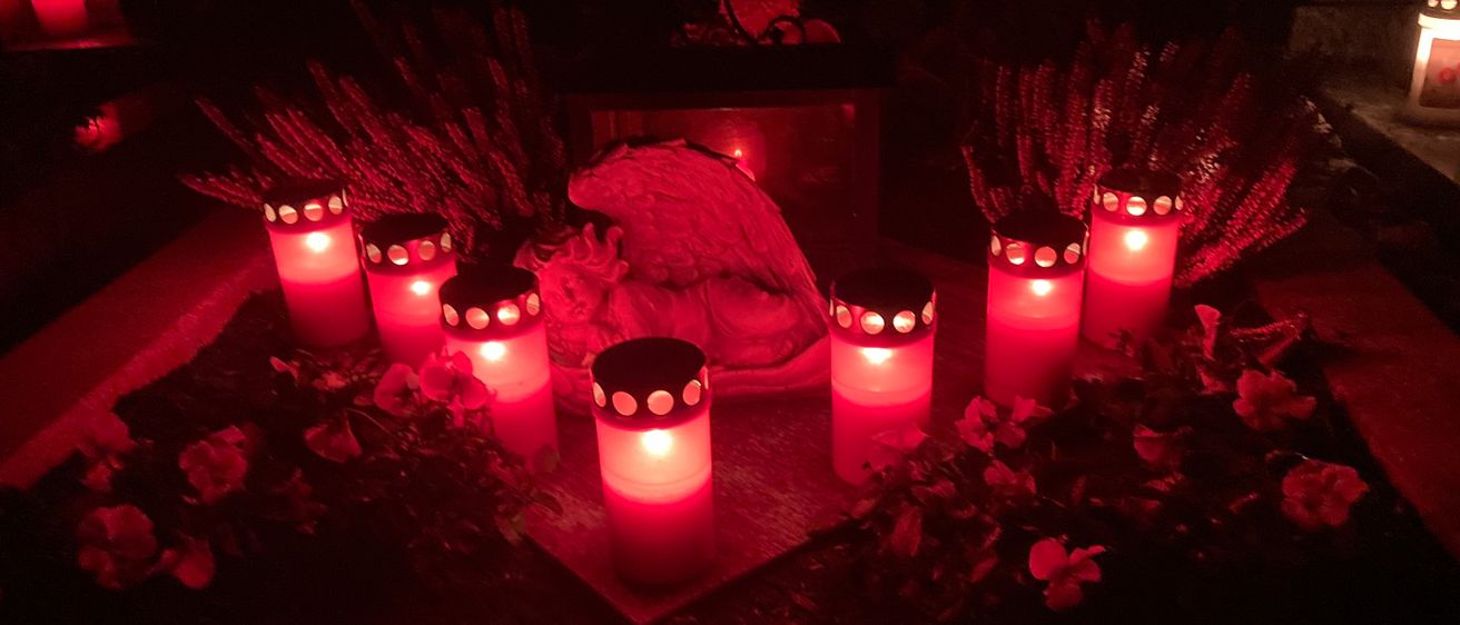 Ein Denkmal mit Kerzen, einer Engelsstatue und Blumen. Die Kerzen sind entzündet und die Engelsstatue ist weiß.