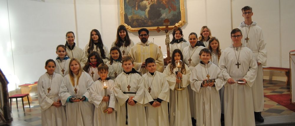 Eine Gruppe von Kindern in weißen Gewändern und mit Kerzen in der Hand posiert für ein Foto in einer Kirche. Ein Priester steht hinter ihnen und ein großes Gemälde ist an der Wand.