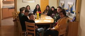 Eine Gruppe von Menschen sitzt um einen Tisch in einem Esszimmer und lächelt für ein Foto. Der Tisch ist mit Blumen, Gläsern und Papieren gedeckt. Eine Lampe hängt von der Decke.