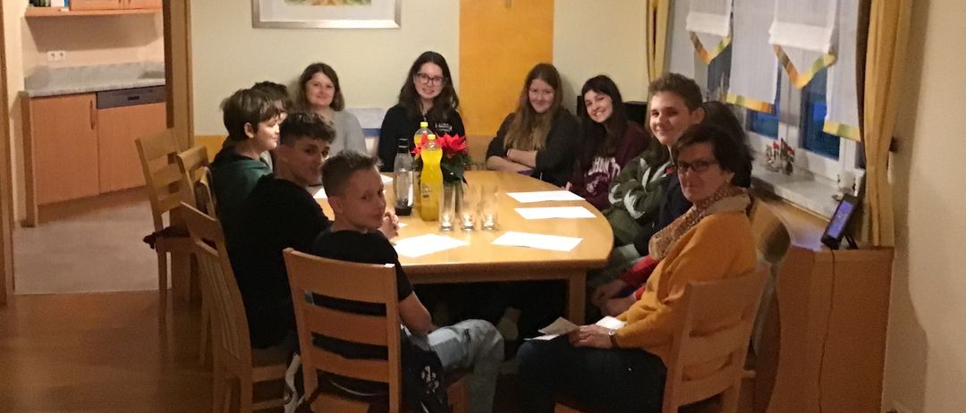 Eine Gruppe von Menschen sitzt um einen Tisch in einem Esszimmer und lächelt für ein Foto. Der Tisch ist mit Blumen, Gläsern und Papieren gedeckt. Eine Lampe hängt von der Decke.