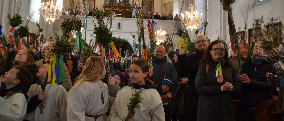 In einer Kirche hält eine Gruppe von Menschen in weißen Gewändern Grünpflanzen und Bänder. Ein Mann und eine Frau in der Front tragen eine Brille. Ein Kind mit einem blauen Hut steht vor ihnen.