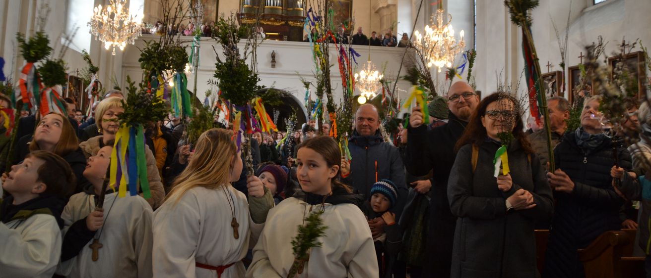 In einer Kirche hält eine Gruppe von Menschen in weißen Gewändern Grünpflanzen und Bänder. Ein Mann und eine Frau in der Front tragen eine Brille. Ein Kind mit einem blauen Hut steht vor ihnen.