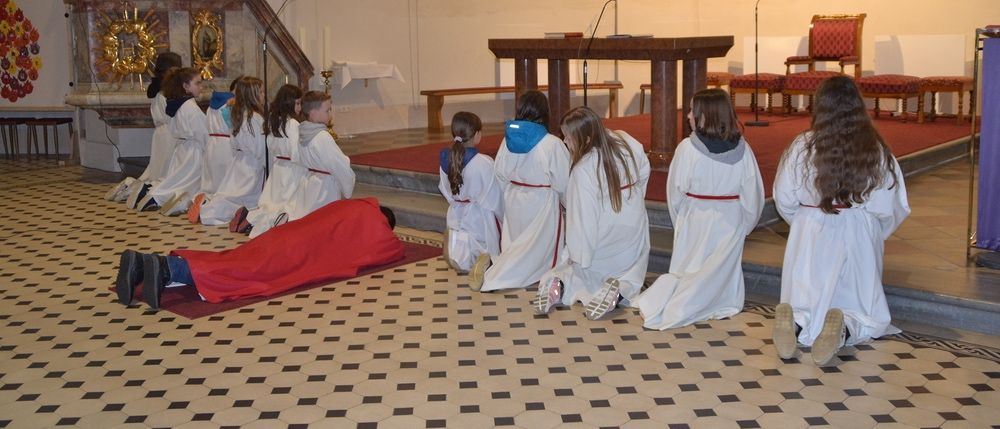 Kinder in weißen Gewändern knien in einer Kirche, eines liegt auf einem roten Tuch. Ein Holztisch mit Mikrofon steht in der Mitte. Ein Gemälde hängt an der Wand.
