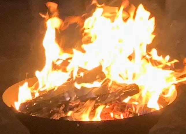 Ein großer Feuerkorb mit brennendem Holz und Flammen, umgeben von Dunkelheit. Das Feuer strahlt ein warmes Licht aus.