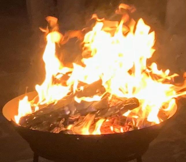 Ein großer Feuerkorb mit brennendem Holz und Flammen, umgeben von Dunkelheit. Das Feuer strahlt ein warmes Licht aus.