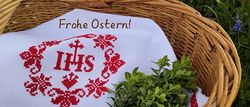 Ein Korb aus Weide enthält ein weißes Tuch mit roter Stickerei, mit dem Text Frohe Ostern! und einem Kreuzsymbol, umgeben von grünen Pflanzen und blauen Blumen.