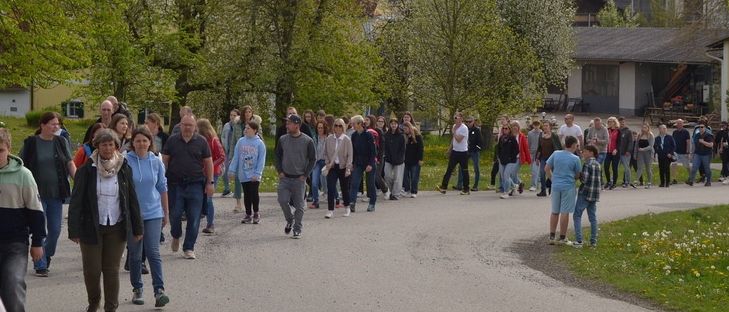 Eine Gruppe von Menschen geht auf einer Straße entlang, die von Bäumen gesäumt ist. Einige tragen Hüte und Turnschuhe, während andere Taschen tragen. Gebäude befinden sich an der Seite.