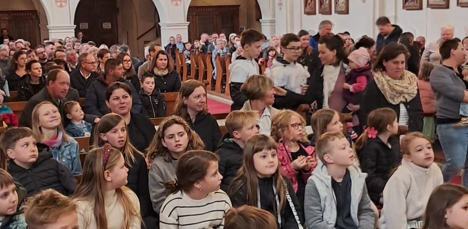 Eine Kirche voller großer Menschenmenge, einschließlich Kinder, sitzend und stehend, mit Kronleuchtern an der Decke und gerahmten Bildern an den Wänden.