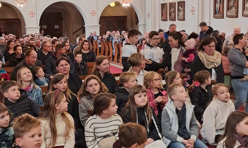Eine Kirche voller großer Menschenmenge, einschließlich Kinder, sitzend und stehend, mit Kronleuchtern an der Decke und gerahmten Bildern an den Wänden.