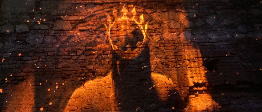 Eine schattenhafte Gestalt mit Krone wird von einem warmen Licht beleuchtet und auf eine Ziegelwand projiziert. Das Licht erzeugt einen flammenartigen Effekt auf dem Kopf der Gestalt.