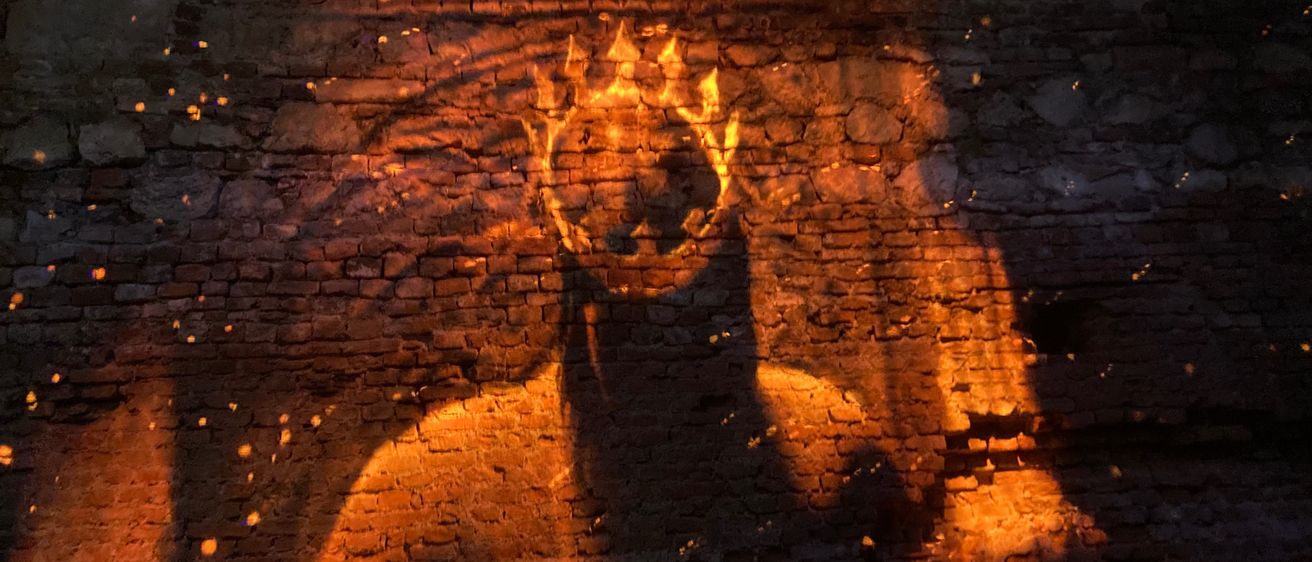 Eine schattenhafte Gestalt mit Krone wird von einem warmen Licht beleuchtet und auf eine Ziegelwand projiziert. Das Licht erzeugt einen flammenartigen Effekt auf dem Kopf der Gestalt.